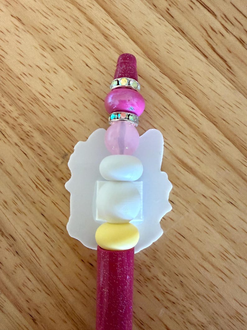 Op de afbeelding: Een pen met een donkerroze glitterhouder, bekroond met een stapel kralen waaronder een donkerroze glitterkraal, twee zilverkleurige strass-tussenstukjes, een lichtroze kraal, twee witte kralen en een gele kraal, met een platte witte uitgesneden vorm.