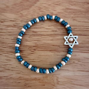 Op de afbeelding: Een kralenarmband met afwisselend teal en crèmekleurige kralen. Het heeft een zilverkleurige Davidster-bedel. De armband wordt getoond op een houten oppervlak. Dit sieraad is geschikt voor dagelijks gebruik.