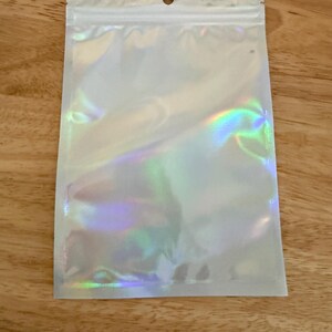 Op de afbeelding: Een rechthoekig, plat, hersluitbaar zakje met een glinsterende, iriserende, holografische afwerking die licht in verschillende kleuren reflecteert. Het bovenste gedeelte bevat een doorzichtige plastic strook met een centraal rond ophanggat.