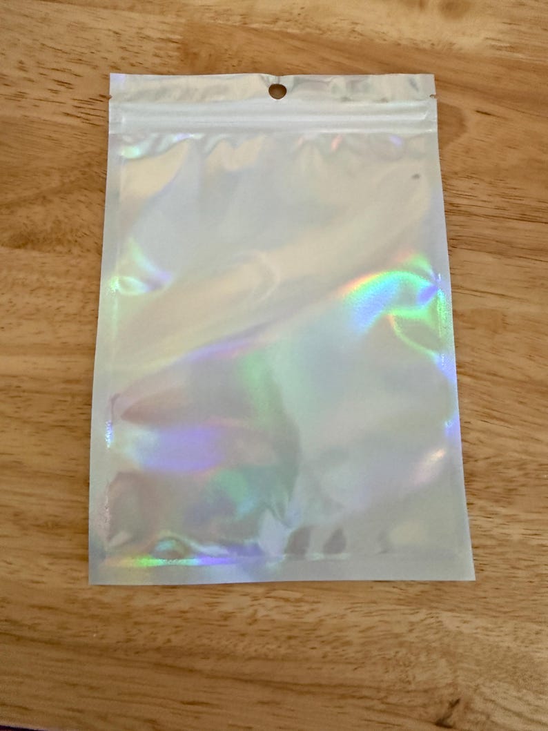 Op de afbeelding: Een rechthoekig, plat, hersluitbaar zakje met een glinsterende, iriserende, holografische afwerking die licht in verschillende kleuren reflecteert. Het bovenste gedeelte bevat een doorzichtige plastic strook met een centraal rond ophanggat.