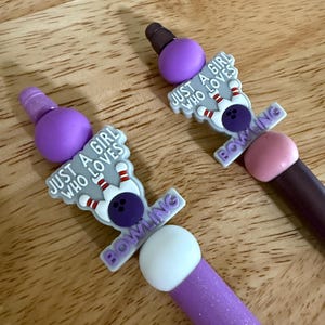 Puede incluir: Dos bolígrafos con adornos temáticos de bolos. Los adornos presentan un diseño de bolos con el texto "JUST A GIRL WHO LOVES BOWLING". Los bolígrafos tienen detalles en morado, rosa y marrón. Los bolígrafos están sobre una superficie de madera.