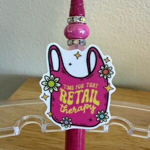 Op de afbeelding: Een magenta glitterpen met een decoratieve topper in de vorm van een roze boodschappentas. De afbeelding bevat gele tekst met de tekst "TIME FOR THAT RETAIL therapy", omringd door kleine witte en gele bloemen en steraccenten.