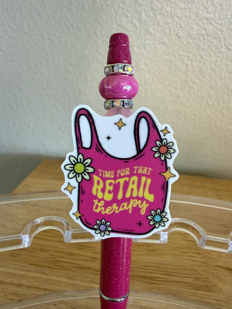 Op de afbeelding: Een magenta glitterpen met een decoratieve topper in de vorm van een roze boodschappentas. De afbeelding bevat gele tekst met de tekst "TIME FOR THAT RETAIL therapy", omringd door kleine witte en gele bloemen en steraccenten.