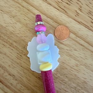 Op de afbeelding: Een glitterende magentakleurige pen met een decoratieve topper in een witte silhouetvorm, geaccentueerd door een stapel kralen in roze, wit en geel, gescheiden door twee zilverkleurige ringen met iriserende kristallen, naast een muntstuk.