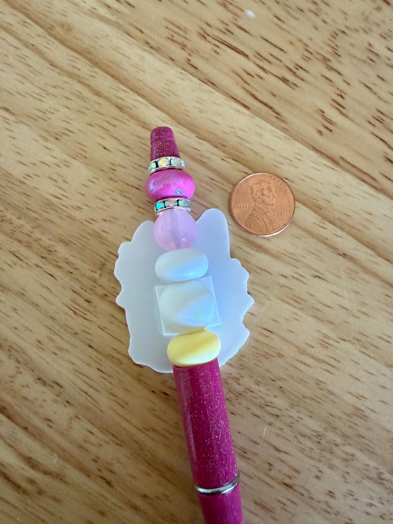 Op de afbeelding: Een glitterende magentakleurige pen met een decoratieve topper in een witte silhouetvorm, geaccentueerd door een stapel kralen in roze, wit en geel, gescheiden door twee zilverkleurige ringen met iriserende kristallen, naast een muntstuk.