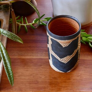 May include: A black and brown ceramic vase with a geometric pattern. The vase is sitting on a wooden table.