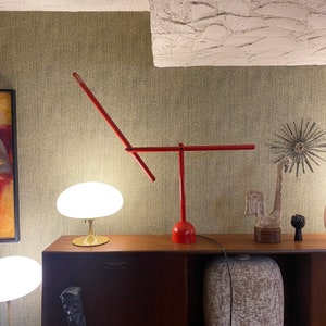 May include: A red desk lamp with a unique, angular design. The lamp has a vertical base and a horizontal arm, with an angled section extending upwards. The lamp is on a wooden surface, with other decorative items in the background.