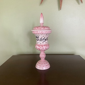 May include: A white ceramic jar with a pink and black floral design. The jar has a lid and is decorated with the word "Rosa" in black lettering.