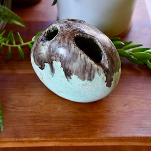 May include: A light green and brown ceramic vase with two holes on the side. The vase is oval shaped and has a speckled glaze.