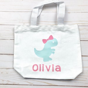 May include: White canvas tote bag with a light blue dinosaur with a pink bow and the name Olivia printed in pink.