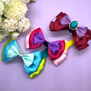 May include: Four colorful satin hair bows in shades of blue, green, pink, purple, and burgundy. One bow features a teal-colored gem. The bows are arranged on a light purple surface with white flowers in the background.
