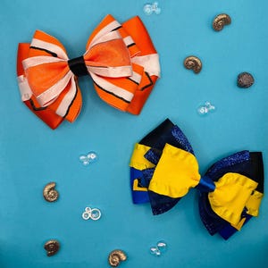 Peut inclure: Deux nœuds pour cheveux faits à la main sur fond bleu. L'un est orange avec des rayures blanches et un centre noir, l'autre est jaune et bleu. Des coquillages et des bulles décoratives sont présents.