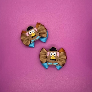 May include: Two handmade hair bows featuring a cartoon character design. Each bow has a brown satin ribbon base, a turquoise accent, and a felt character face with a black hat, pink cheeks, and a yellow nose. The background is a solid magenta color.