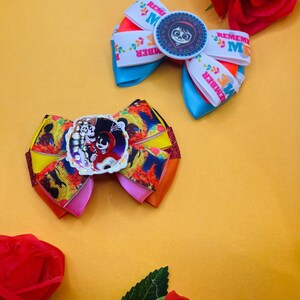 May include: Two vibrant hair bows with a Coco theme. One bow features a round emblem with a character's face, and the other has a similar design. The bows are made of various ribbons in shades of blue, orange, pink, and yellow.