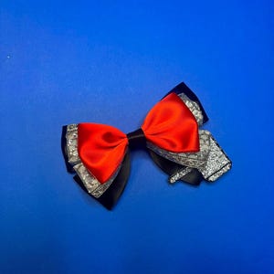 Pode incluir: Um laço vermelho com detalhes em preto e prata. O laço é feito de cetim e tem um acabamento brilhante.