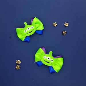 May include: Two green alien-themed hair bows with three eyes, green and blue ribbons, and gold star-shaped embellishments on a blue background. These bows are likely for children.