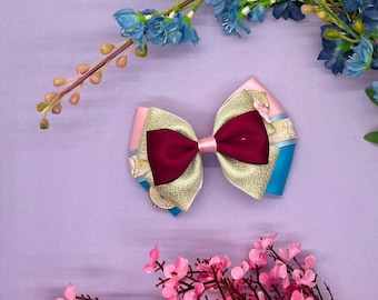 May include: A decorative hair bow featuring a burgundy center, gold glitter accents, and pink and blue ribbons. The bow is set against a purple background with floral accents, creating a visually appealing accessory.