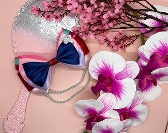 May include: A blue, pink, and red ribbon bow with a white pearl accent. The bow is on a silver and white hand mirror with a pink background. The mirror is surrounded by pink flowers.