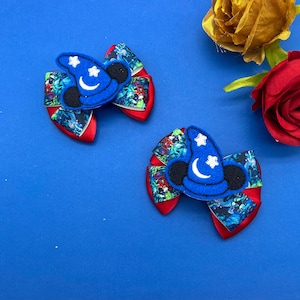 May include: Two blue and red hair bows with a wizard hat design. The bows have a red base with a colorful patterned overlay and a blue wizard hat with white stars and a crescent moon.