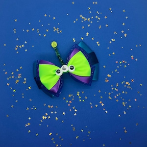 May include: A green, purple and blue bow with three googly eyes. The bow is on a blue background with gold glitter.