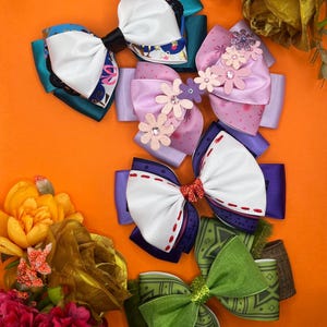 May include: An assortment of colorful hair bows in various designs. The bows feature white, blue, purple, and green ribbons, some with floral embellishments and glitter accents. The bows are arranged on an orange surface.