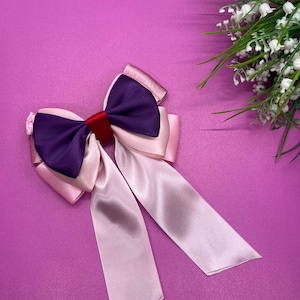 Pode incluir: Um laço de cabelo grande e feito à mão com um design em camadas. Possui um centro roxo escuro, detalhes em rosa claro e fitas longas e esvoaçantes. O laço é preso com uma pequena faixa vermelha e fica em um fundo roxo sólido. Um raminho de flores brancas está ao fundo.
