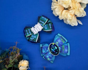 May include: Two hair bows with a blue and green color scheme. One bow features a white graphic of a group of figures, the other features a blue and green glitter fabric with a circular graphic of a group of figures.