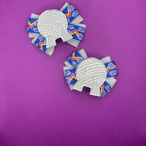 May include: Two handmade hair bows featuring a silver, glittery sphere design with a gray base. The bows have a blue ribbon with the text "EPCOT CENTER" in white. The background is a solid purple.