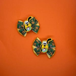 May include: Two yellow and green hair bows featuring a cartoon dog character. The bows have a yellow border and a green center with a repeating pattern. The cartoon dog is yellow with black accents and a green tongue.
