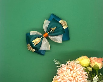 May include: A teal blue bow with a gold arrow accent. The bow is made of satin and tulle and has a gold trim.