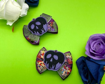 Pode incluir: Duas presilhas de cabelo em forma de maçã roxa e preta com um design de caveira, sobre um fundo verde vibrante. As presilhas são decoradas com fitas com vilões da Disney. Flores decorativas brancas, azuis e roxas circundam as presilhas.