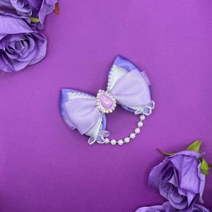 Puede incluir: Un lazo para el cabello lavanda con una joya central y una cadena de perlas. El lazo está rodeado de rosas moradas sobre un fondo morado a juego. El lazo presenta un acabado satinado y un ribete de encaje blanco.