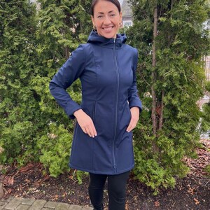 Könnte beinhalten: Eine marineblaue Kapuzenjacke mit durchgehendem Reißverschluss und Taschen. Die Jacke wird mit schwarzen Leggings und schwarzen Stiefeln getragen. Die Person steht vor grünen Bäumen.