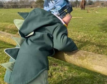 May include: A child wearing a dark green dinosaur-themed hooded jacket with white and light green accents. The jacket has a row of triangular spines down the back. The child is leaning on a wooden fence in a grassy field.