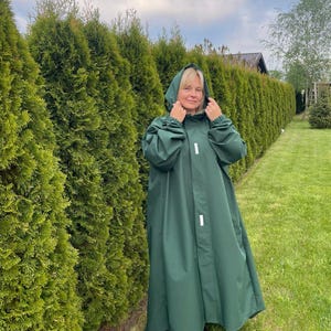 May include: A long, dark green hooded raincoat featuring a front placket with two visible white rectangular reflective strips. The garment has long sleeves with elasticized cuffs and a loose, flowing silhouette that extends to the lower legs.