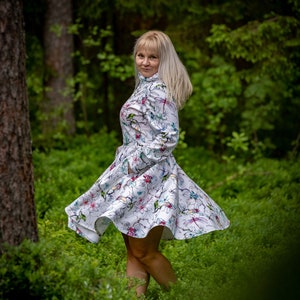Puede incluir: Un vestido blanco con estampado floral y de libélulas. El vestido tiene mangas largas y una falda acampanada. La mujer está de pie en un bosque con follaje verde y árboles al fondo.