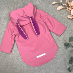 Puede incluir: Chaqueta con capucha rosa con orejas de conejo adjuntas. Las orejas son moradas con ribete blanco. La chaqueta tiene mangas tres cuartos y un dobladillo curvo. Un parche rectangular blanco está cosido en la parte trasera de la chaqueta.