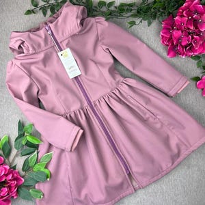 Puede incluir: Chaqueta softshell rosa empolvado con capucha y cremallera morada de largo completo. La chaqueta presenta un diseño de falda acampanada y mangas largas. Una etiqueta está adherida a la cremallera. La chaqueta se exhibe sobre una superficie gris con detalles florales.