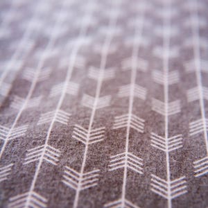 May include: Close-up of a grey fabric with a white arrow pattern. The arrows are arranged in a repeating pattern, pointing in the same direction.