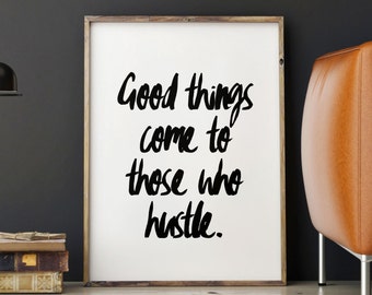 May include: A rectangular wooden frame containing a white print with the black handwritten text Good things come to those who hustle. The frame is positioned upright on a light-colored wooden floor next to a stack of books and a black desk lamp.