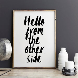 May include: A rectangular framed wall art piece with a light wood frame and a white background featuring the black hand-lettered text "Hello from the other side" in a vertical orientation. The print is displayed on a wooden surface.