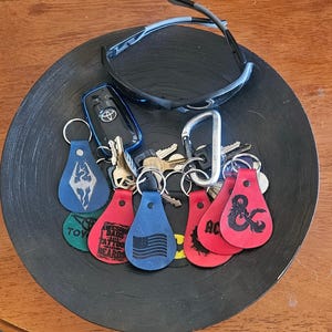 May include: A collection of keys, keychains, and sunglasses in a black vinyl record bowl. Keychains are blue and red, with various designs including a flag, a logo, and text. The sunglasses are black and gray.