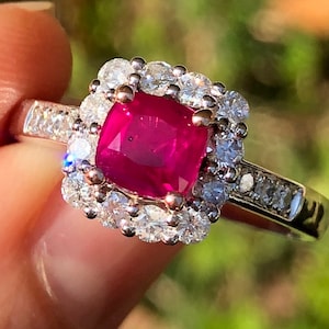 Puede incluir: Un anillo de plata con un gran rubí rojo cuadrado rodeado de diamantes más pequeños. El anillo tiene una banda de diamantes más pequeños.