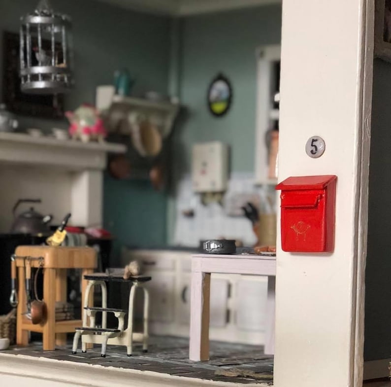 Op de afbeelding: Een rode miniatuur brievenbus van metaal met een klein goudkleurig embleem op de voorkant, gemonteerd op een wit deurkozijn naast een rond zilverkleurig plaatje met het nummer 5. De brievenbus bevindt zich voor een gedetailleerde poppenhuiskeuken.