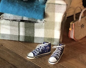 Op de afbeelding: Een paar miniatuur blauwe hoge sneakers met witte veters, witte rubberen neuzen en zwarte randen langs de zolen. De schoenen hebben een klein rond logo aan de zijkant en staan voor een fauteuil met ruitpatroon en een kleine handtas.