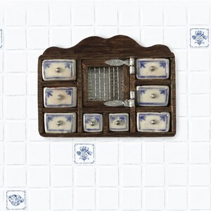 Op de afbeelding: Een miniatuur houten kruidenkastje voor aan de muur met een donkerbruine afwerking, een centrale deur van gaas met zilveren scharnieren en zeven keramische lades. Elke lade is wit met blauwe bloemmotieven en een kleine zilveren knop.