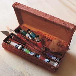 Puede incluir: Una caja rectangular de textura marrón que contiene varios artículos de arte pequeños, incluyendo varios tubos de pintura en verde, blanco y rojo, una espátula de metal, una herramienta con mango de madera y una pequeña esponja beige en el compartimento.