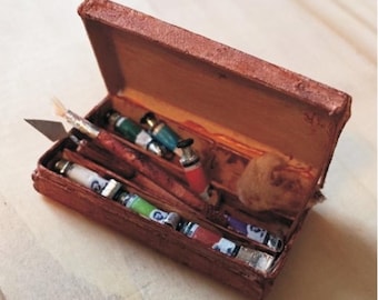 Op de afbeelding: Een rechthoekige bruine doos met een textuur die diverse kleine kunstbenodigdheden bevat, waaronder verschillende verftubes in groen, wit en rood, een metalen paletmes, een gereedschap met houten handvat en een klein beige sponsje in het vak.