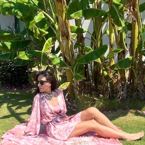 May include: A woman wearing a pink tie-dye robe and matching dress, seated on a pink blanket on a grassy lawn. She is wearing sunglasses and a silver necklace. The background features banana trees and sunlight.
