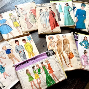 May include: A collection of vintage sewing patterns featuring illustrations of clothing designs for women, men, and children. The patterns showcase various styles, including dresses, shirts, and pajamas. The covers display colorful drawings and text.